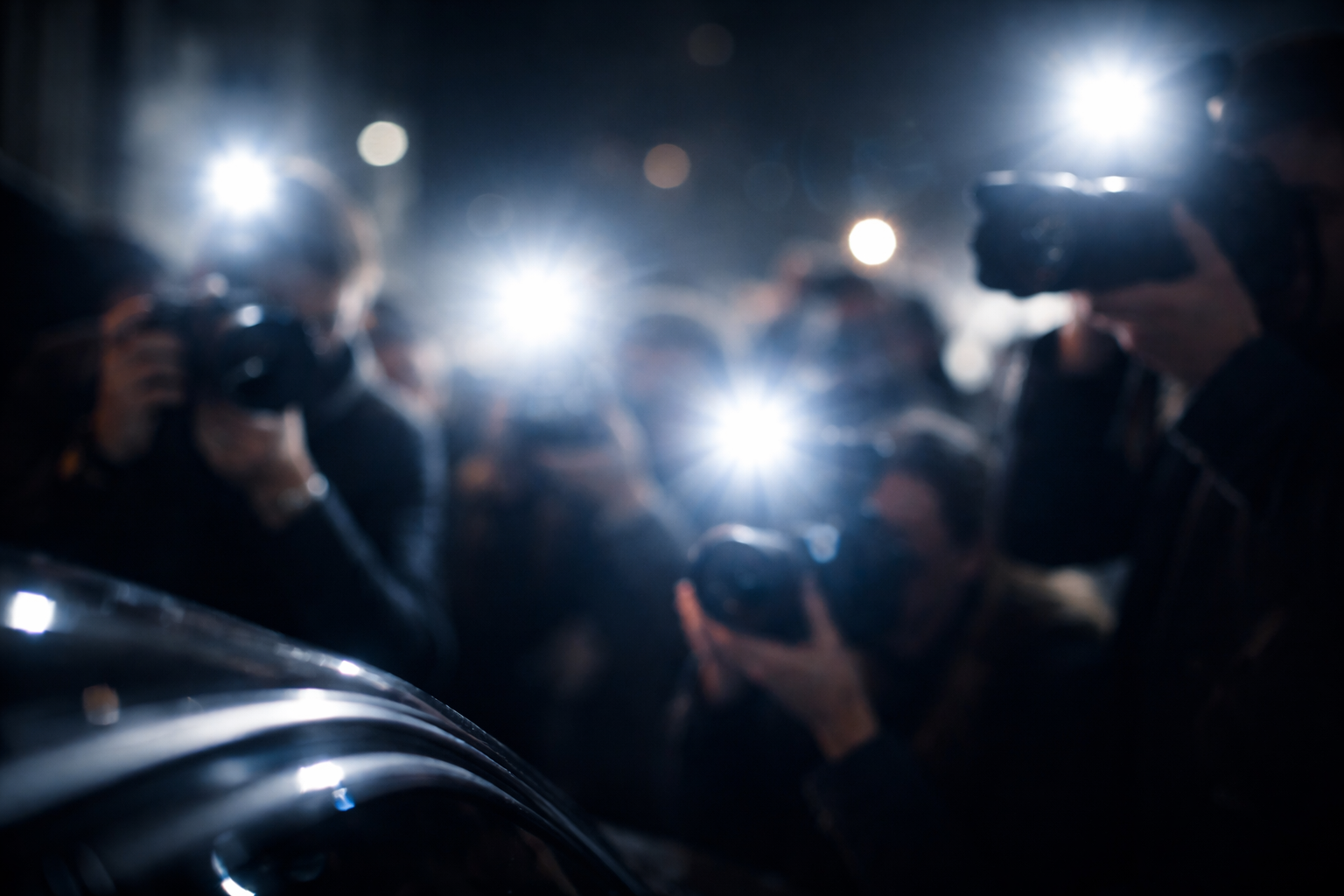 A blurred crowd of photographers at night, taking photos with bright, synchronized camera flashes. A dark, shiny car is visible in the foreground.