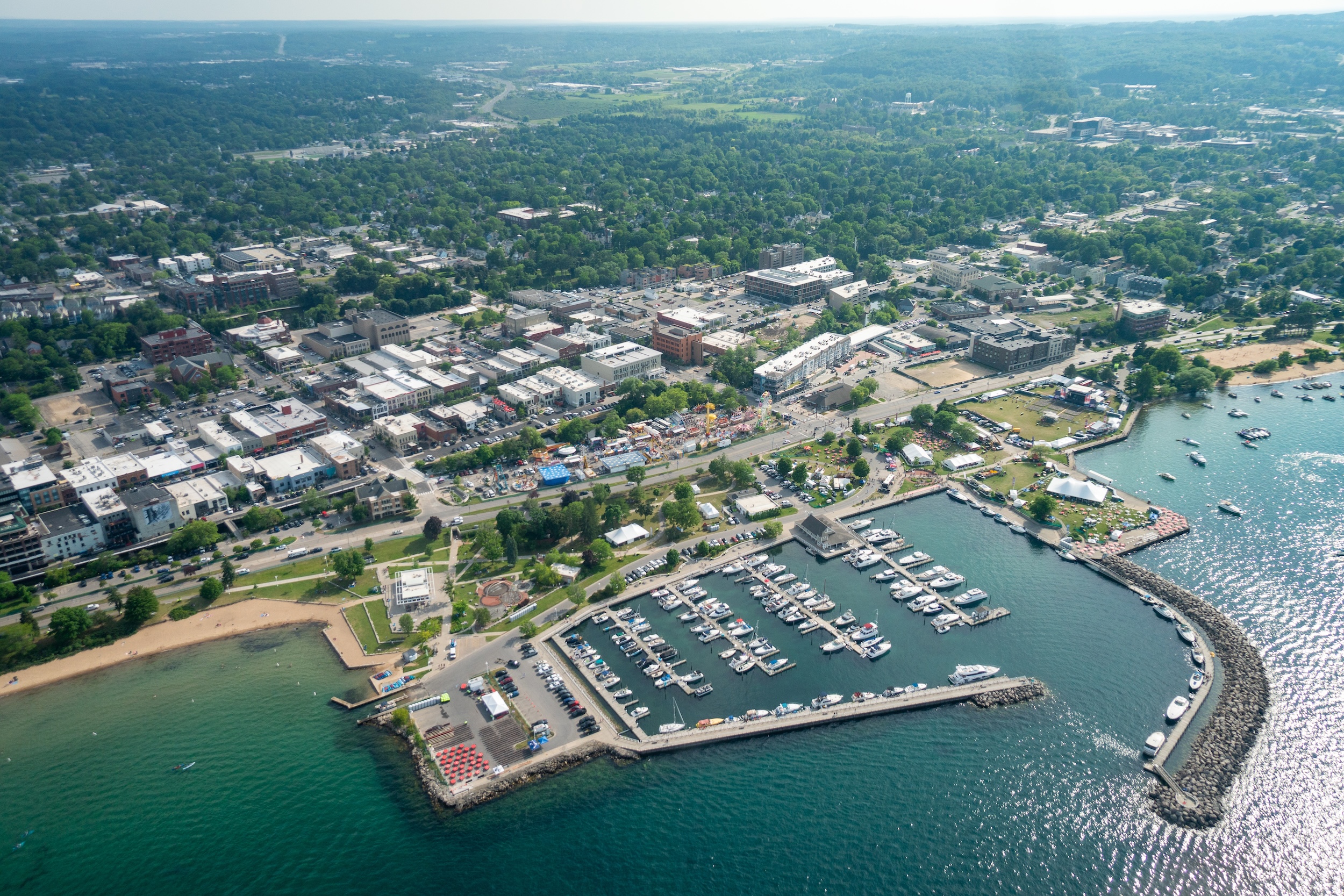 An aerial view of Traverse City's downtown and waterfront. The bright blue harbor holds a busy marina filled with boats, backed by commercial buildings and dense green trees.