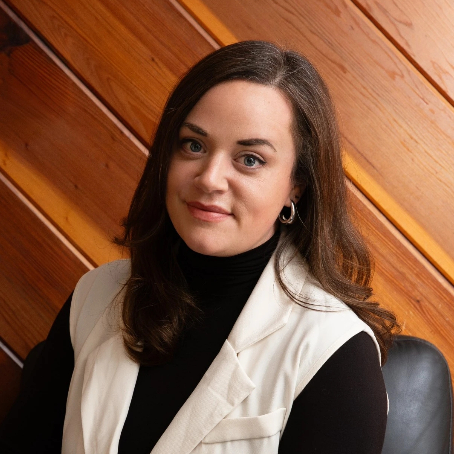 Bevin Buchler smiles professionally in a white vest and black top, standing in front of a warm, diagonally oriented wooden wall.