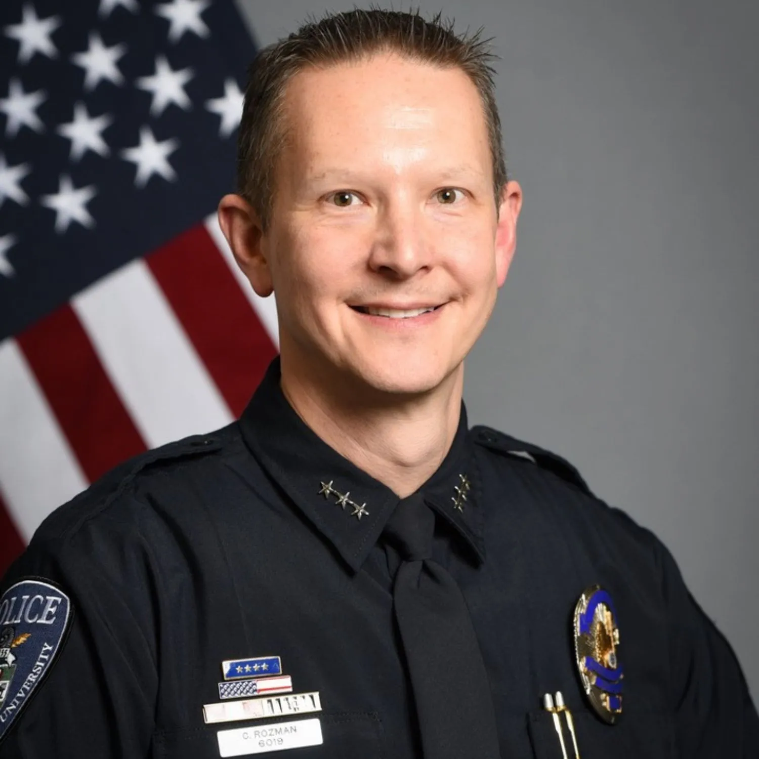 Chris Rozman, a police commander, smiles in his dark uniform with three rank stars and service ribbons, standing before an American flag.