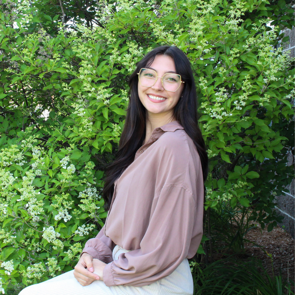 Jenna Baker wearing a mauve shirt and gold-rimmed glasses smiles warmly at the camera while sitting outside in front of a bright green flowering hedge.