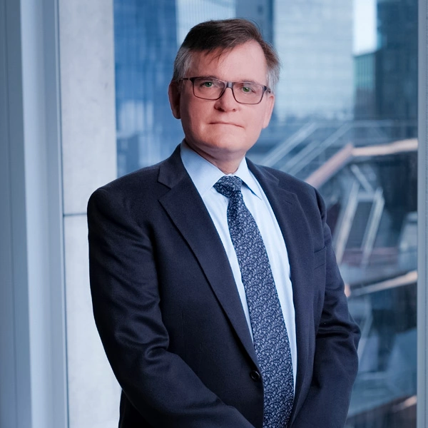 James R. Marsh, wearing a dark suit and glasses, stands in an office in front of a window overlooking a metropolitan skyline.