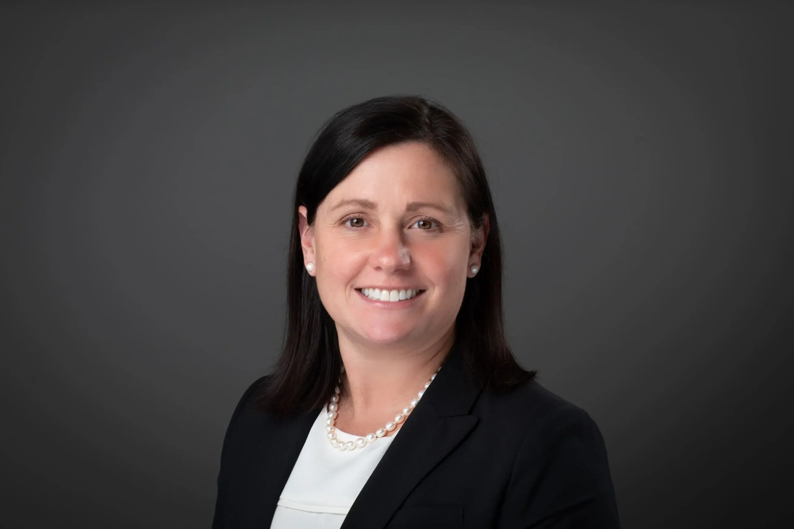 Noelle Moeggenberg, a smiling professional woman with dark hair wears a black suit, a white top, and a pearl necklace against a dark background.