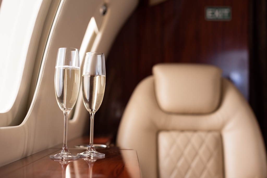 Two champagne flutes on a wooden table in a private aircraft next to a window and a luxurious beige leather seat.