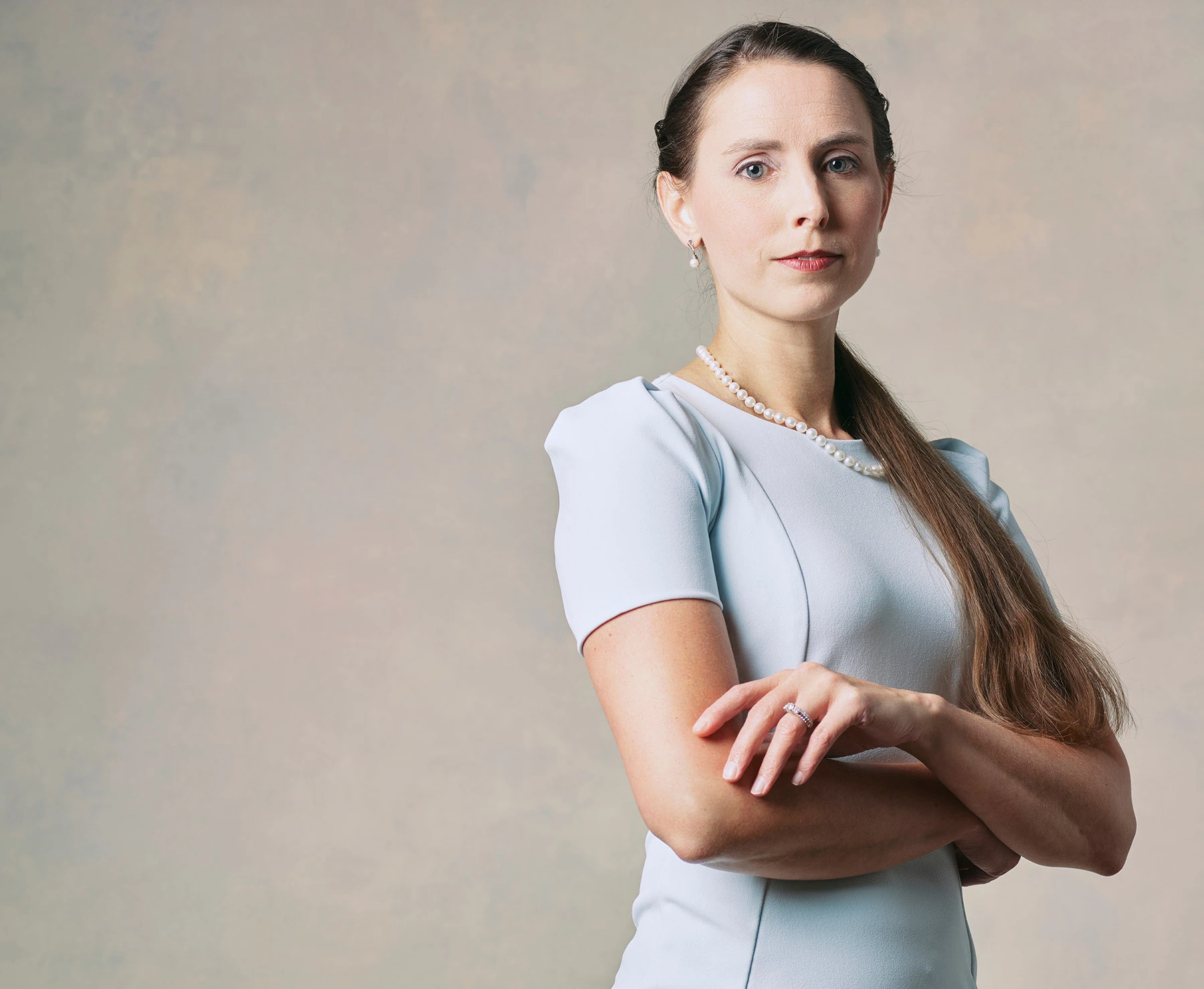 Rachael Denhollander, a female attorney in a light blue dress and pearl jewelry stands with her arms crossed, looking directly at the camera with a confident expression.