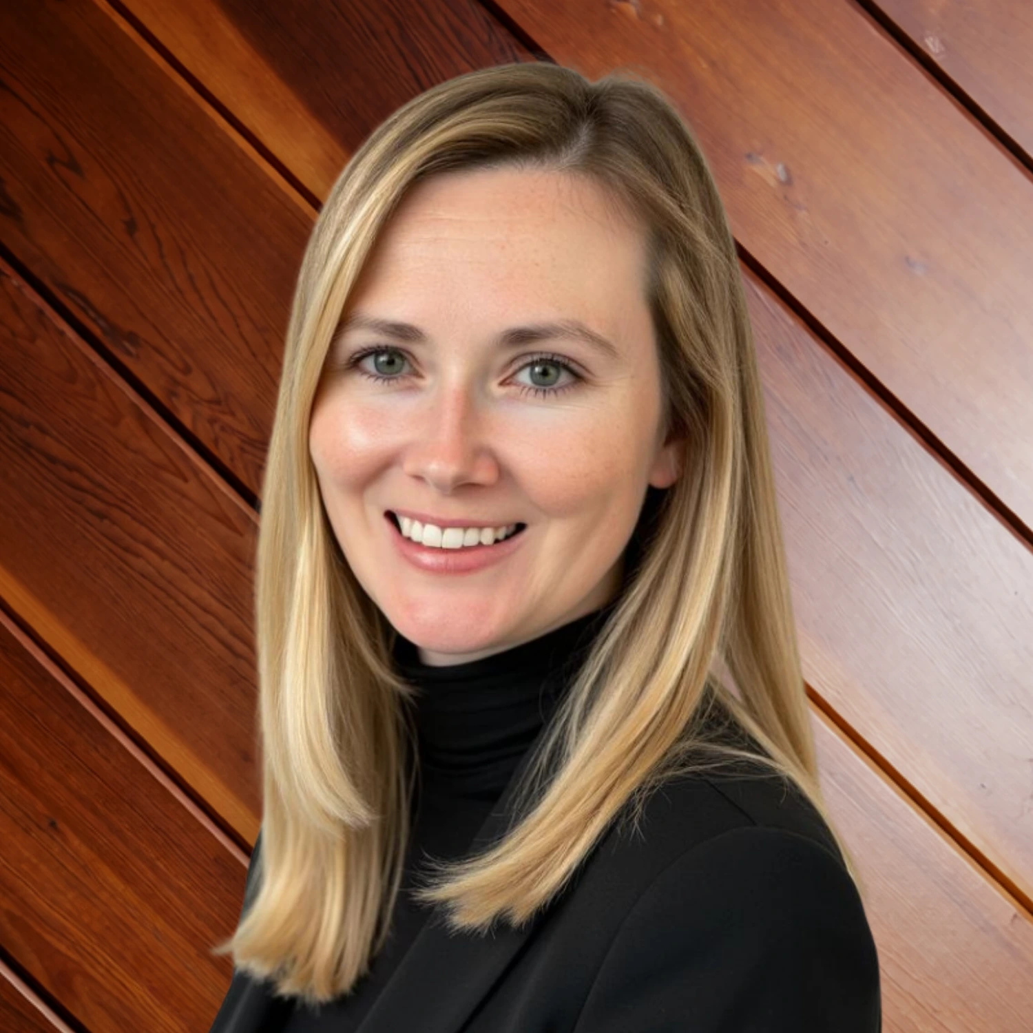 Caitlin McCartney warmly, wearing a black blazer and turtleneck top, standing in front of a warm, diagonal wood plank wall.