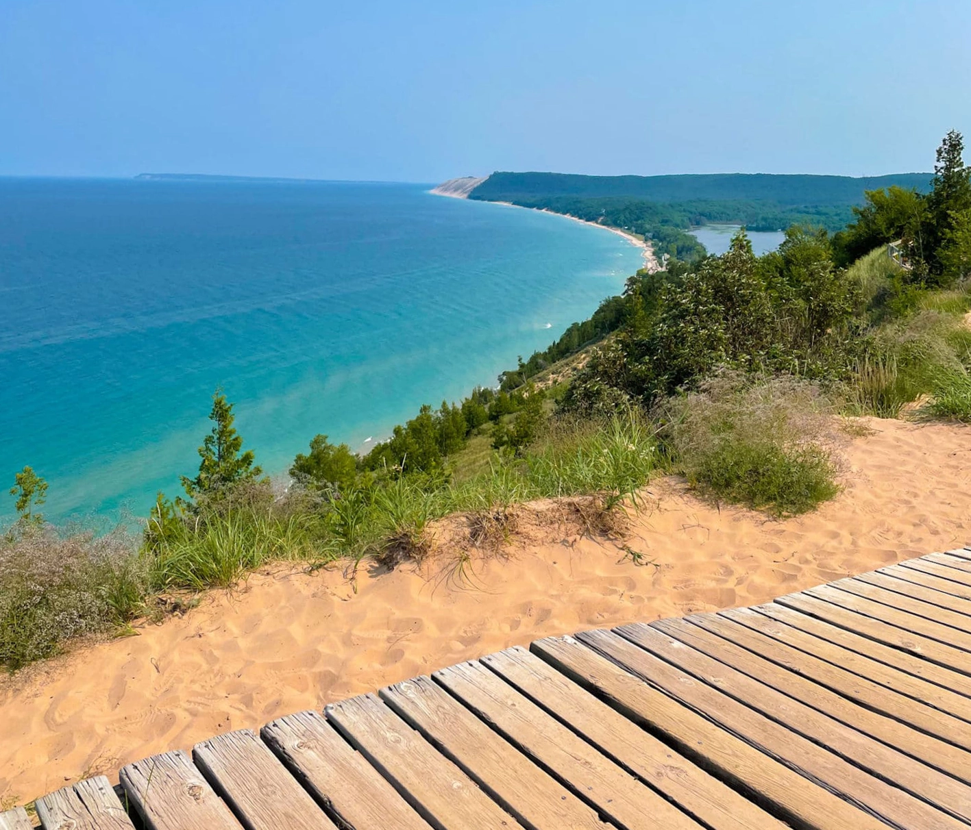 A view from a wooden deck overlooking a steep Lake Michigan sand dune, turquoise water, a forested coastline, and large sand dunes in the distance.