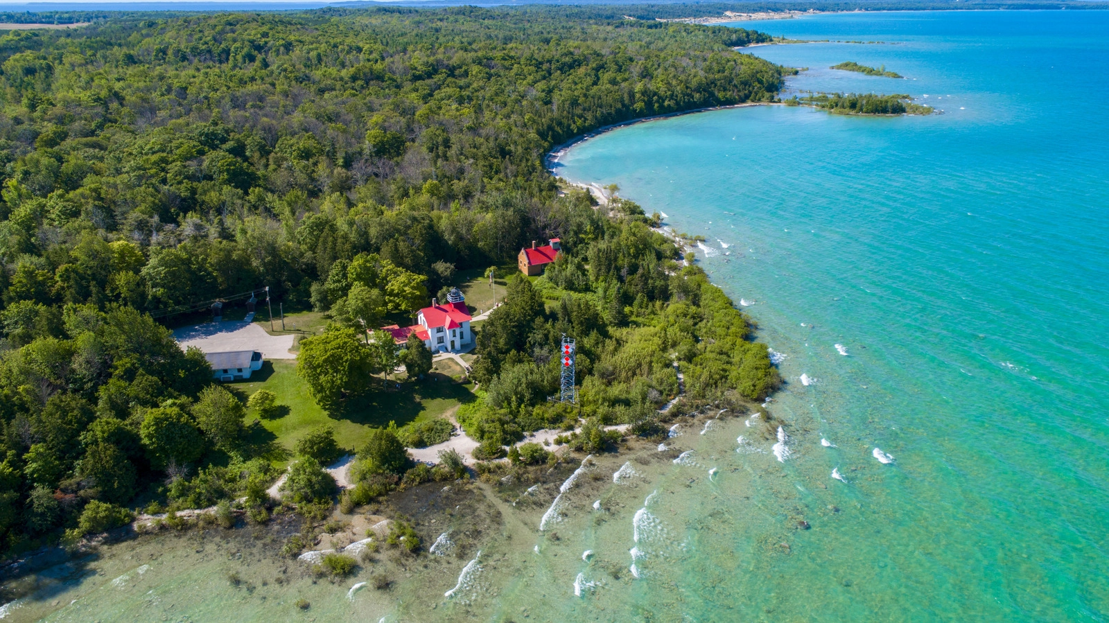 Aerial view of a white lighthouse and outbuildings with red roofs, surrounded by dense green forest on a point of land next to clear, vivid turquoise water.