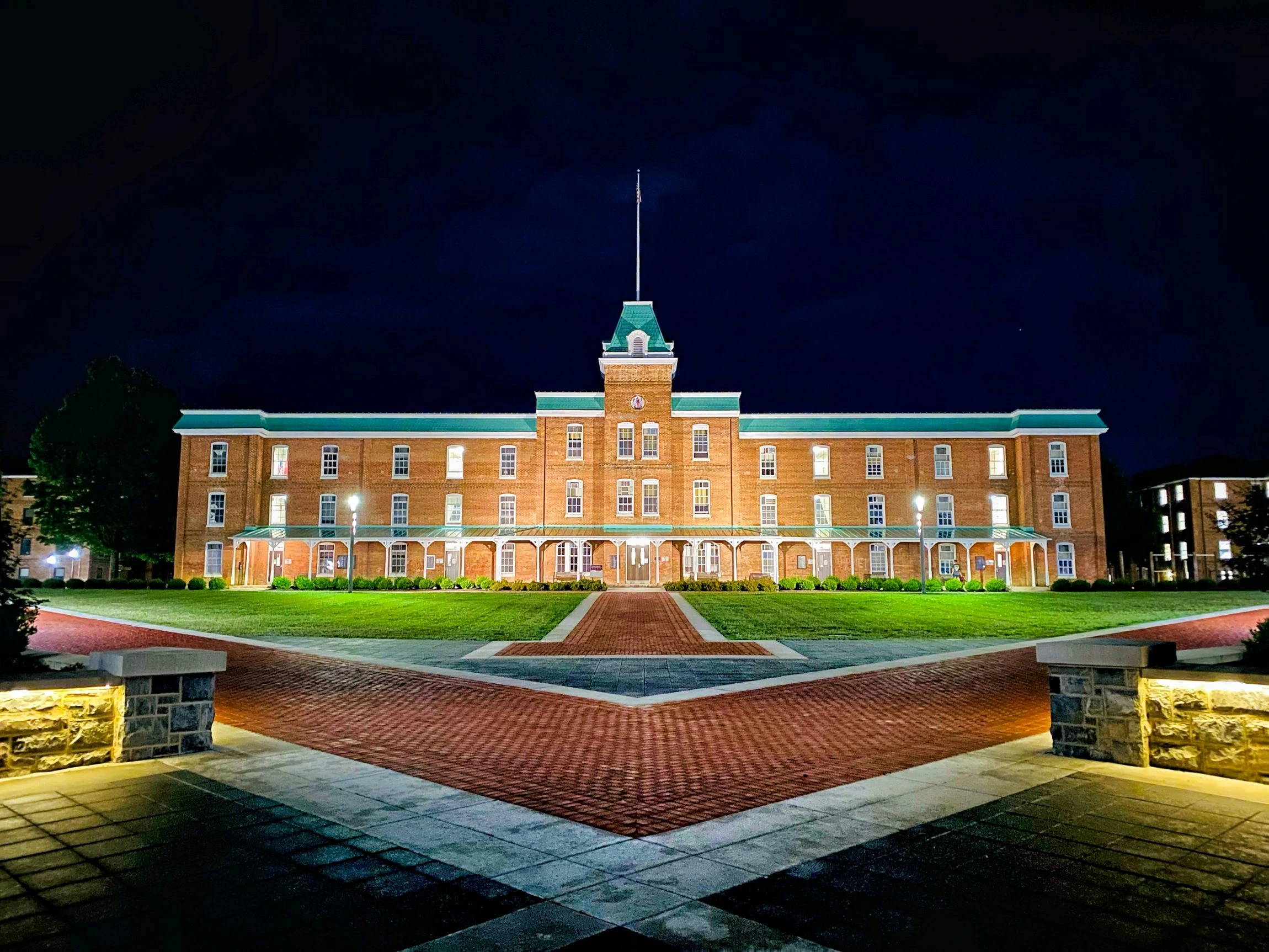 A large, historic brick college building illuminated at night. A long, symmetrical brick walkway leads across the manicured lawn to the central entrance.