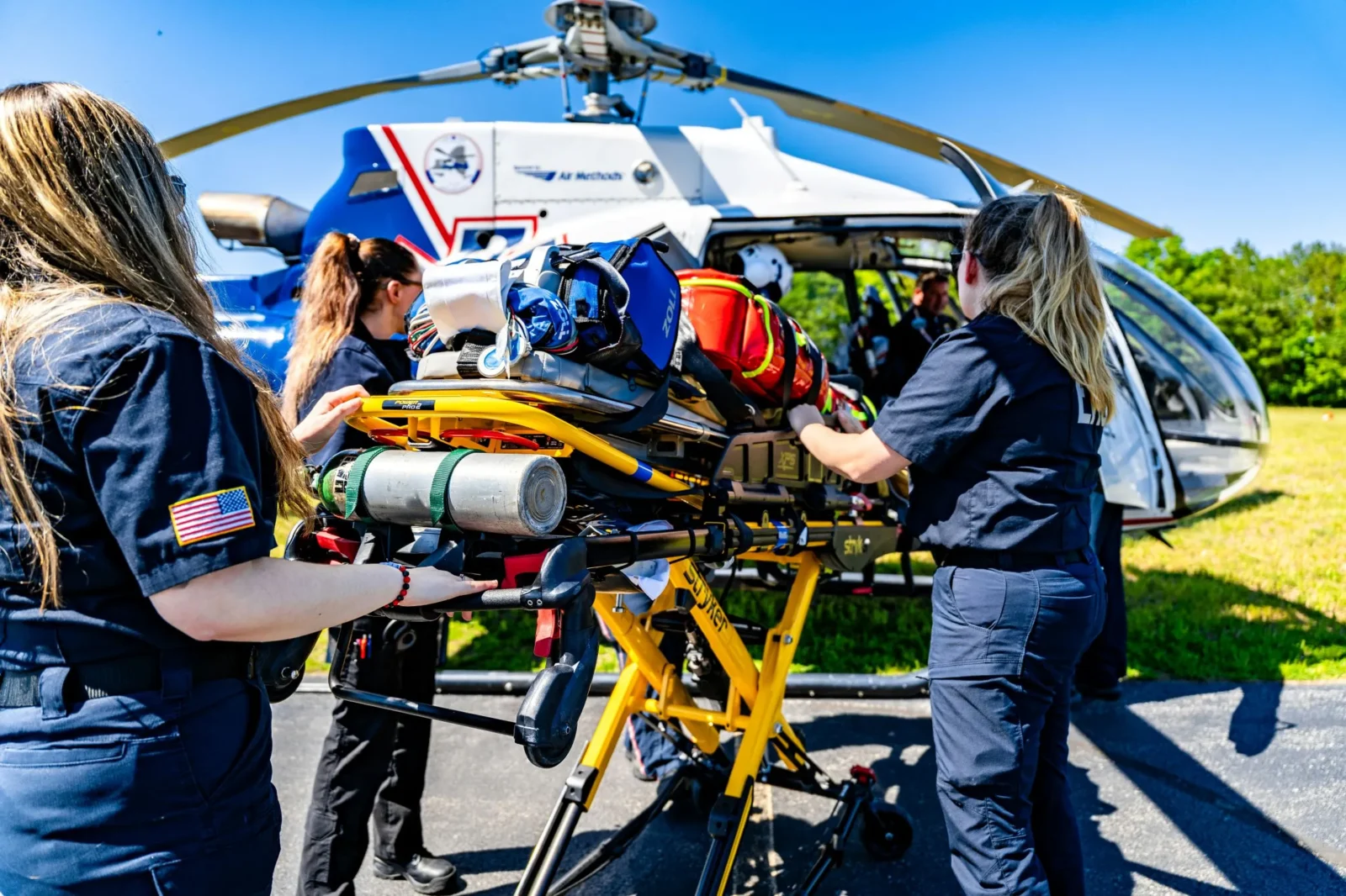 Members of an emergency flight crew load a stretcher equipped with essential medical bags and oxygen tanks into a rescue helicopter on a bright, clear day.