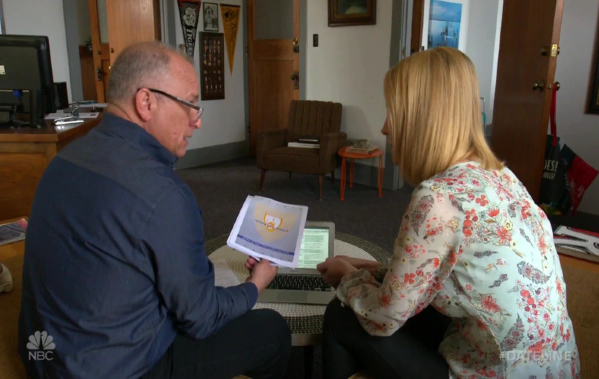 A male attorney with glasses and a blue shirt sits with a blonde female client, reviewing documents and a laptop screen in a room with academic pennants.