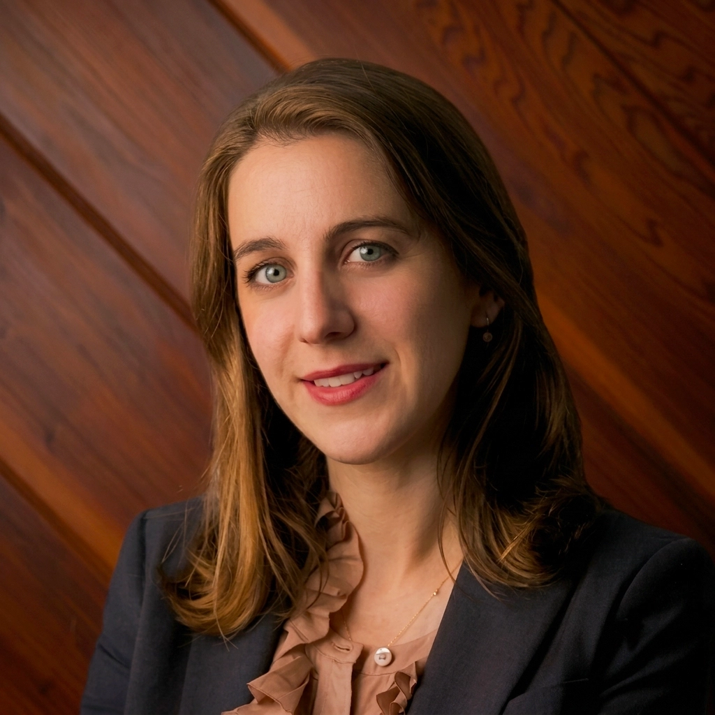 Olympias Iliana Konidaris, an attorney, smiles in a professional headshot. She wears a dark blazer over a ruffled blouse against a warm wood paneled background.