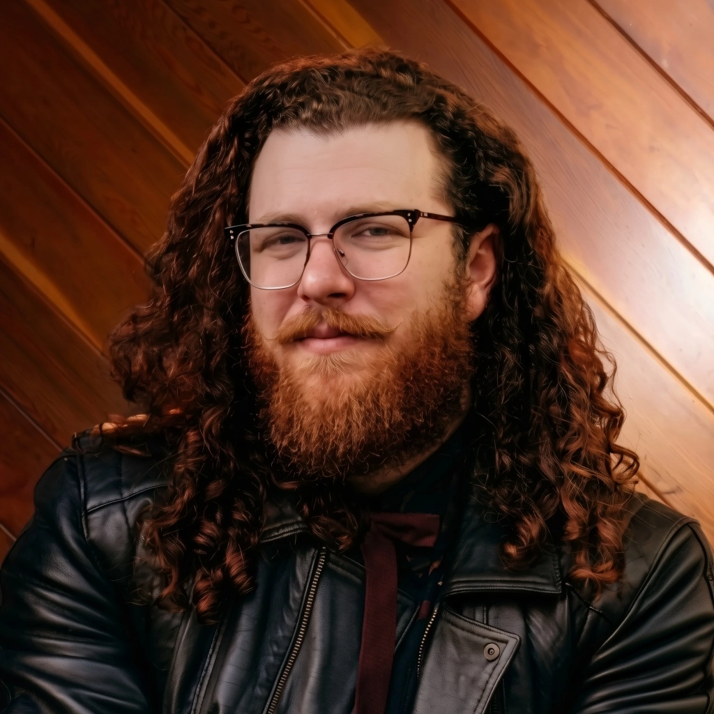 Jacob, a male bodied person with long curly hair, a reddish beard, and glasses smiles confidently. They wear a black leather jacket and a dark tie against a diagonal wooden wall.