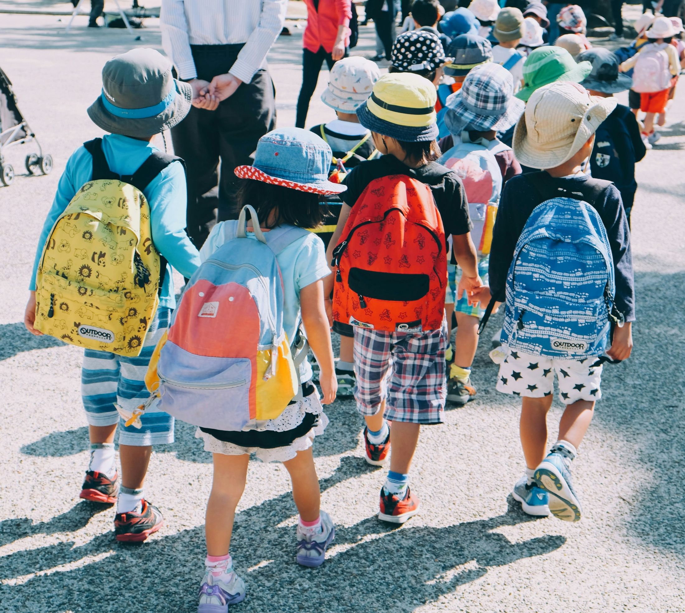 A group of young children wearing colorful hats and backpacks walk together outdoors on a sunny day. They are being guided by adults in the background.