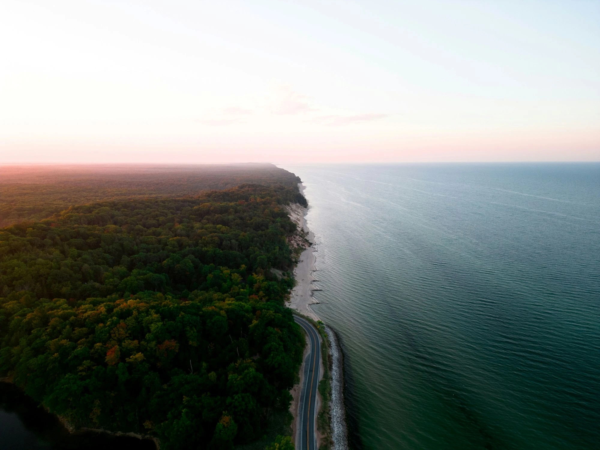 An aerial view of a road winding along the shore between a dense green forest and a wide expanse of deep blue water, illuminated by the pink light of twilight.