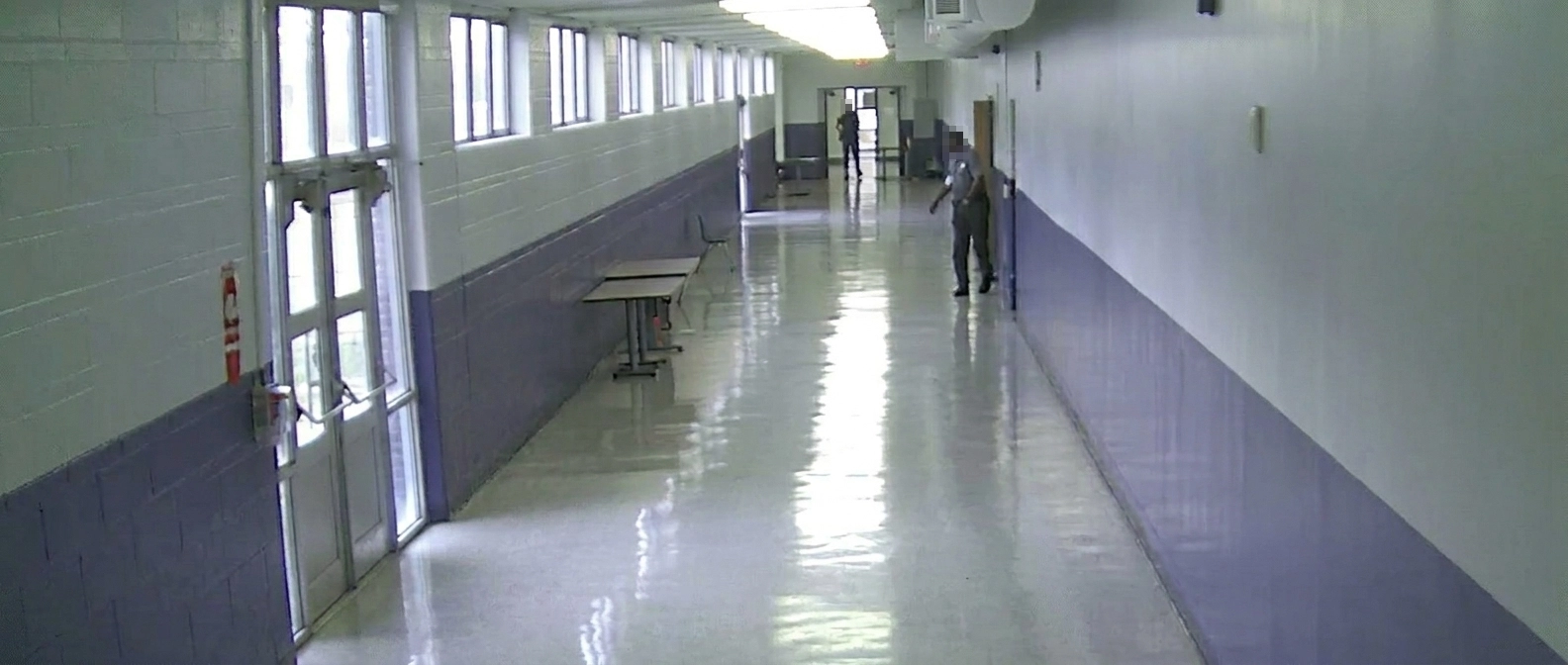 A long, brightly lit school hallway with polished floors and white cinder block walls accented with blue. School shooter enters school confronting staff member