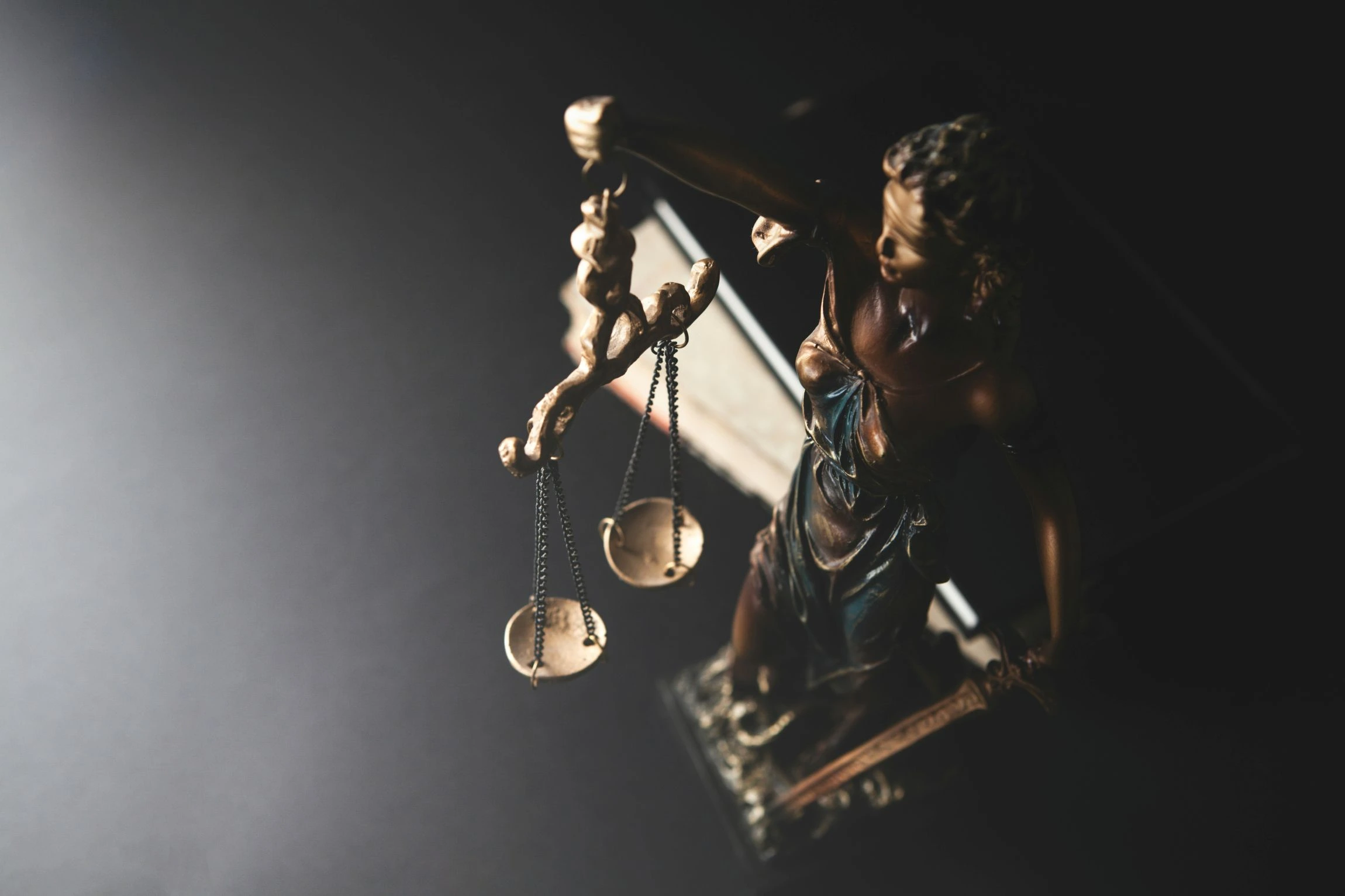 A close-up view of the bronze Lady Justice statue holding the scales of justice, dramatically highlighted by a beam of light in a dark environment.