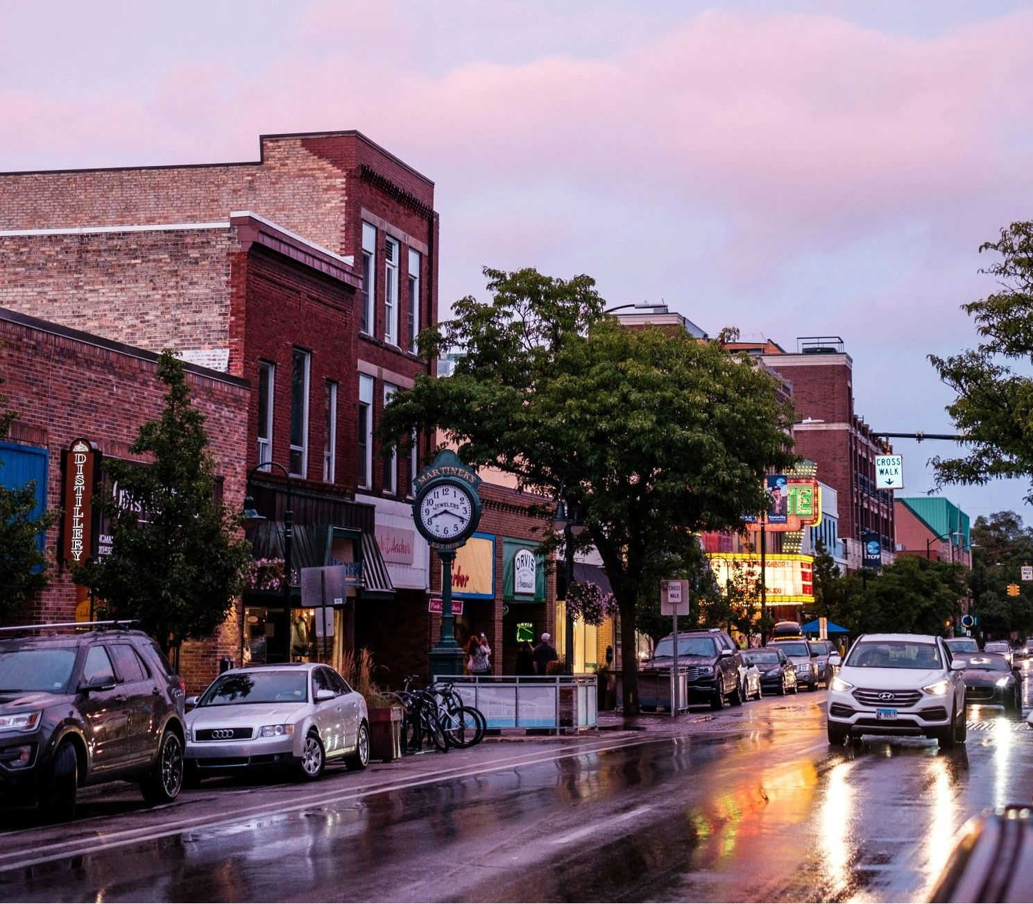 A vibrant urban street scene at dusk. The wet asphalt reflects the warm glow of the neon signs lining the historic brick buildings and storefronts.