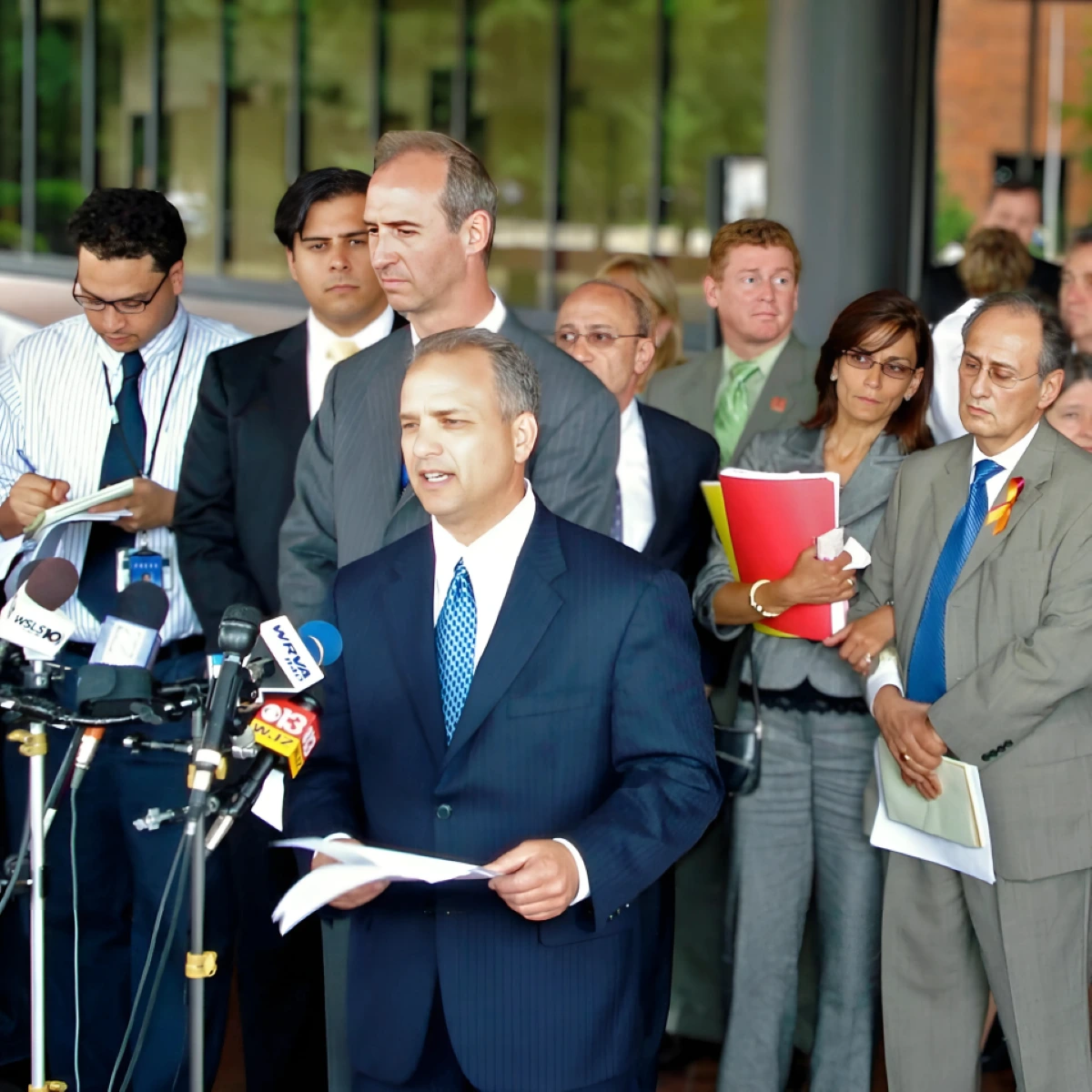 A professional man in a dark suit gives a serious statement, speaking into multiple microphones at a press conference. Attentive colleagues stand behind him.