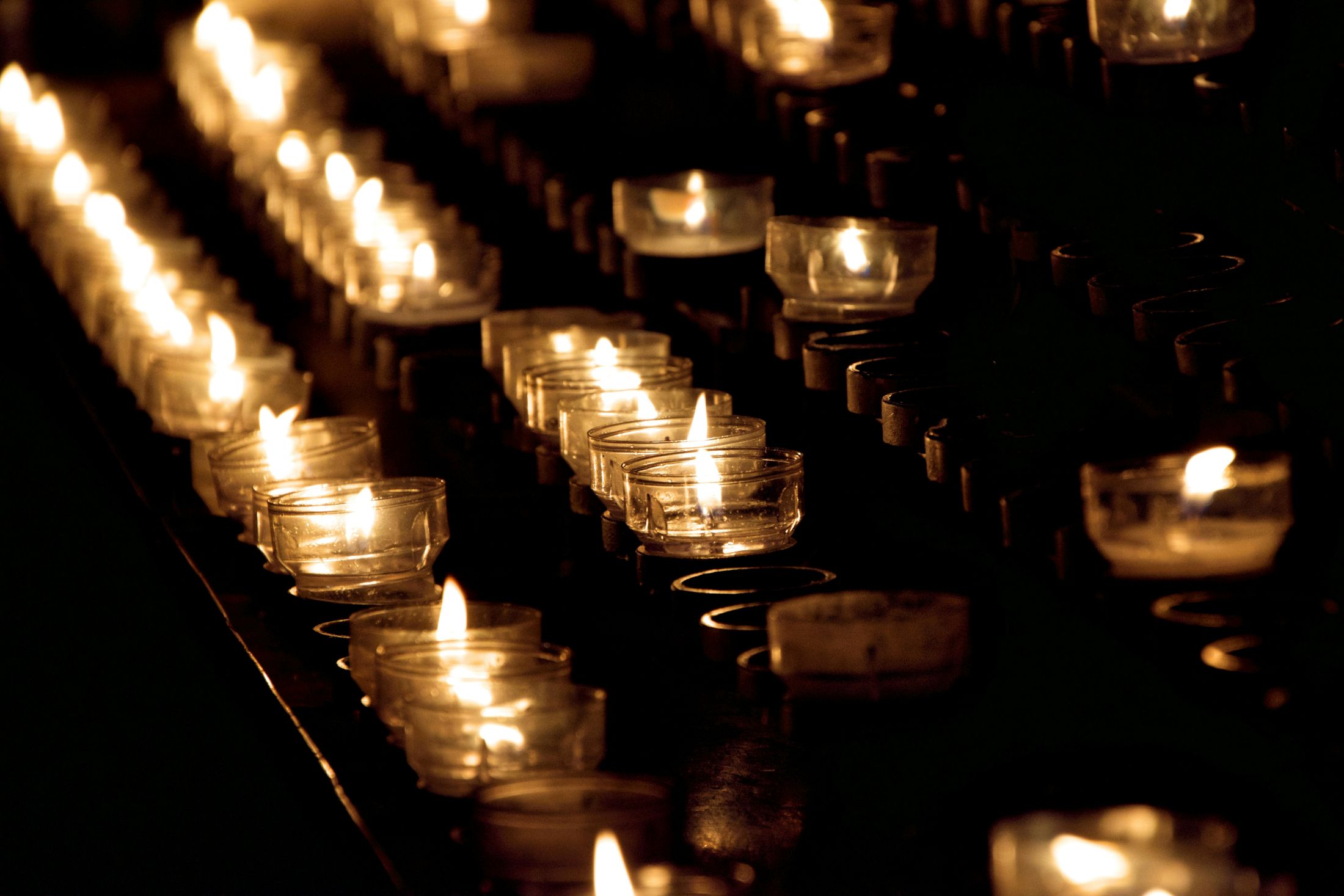 A close-up view of several rows of small, lit candles in clear glass holders, their flames casting a warm, serene glow in the dark.