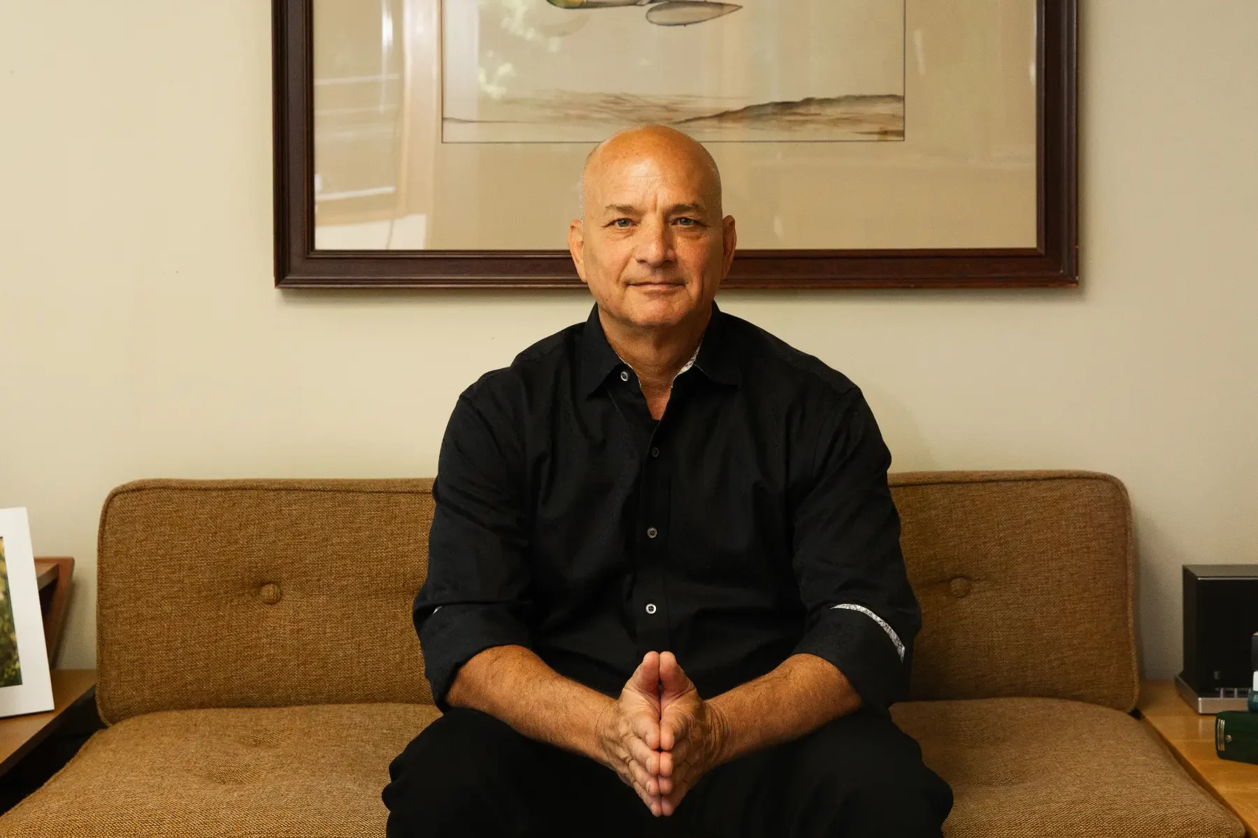 Attorney Doug Fierberg sits on a brown couch with a serious expression, hands clasped. A framed military airplane print hangs above him.