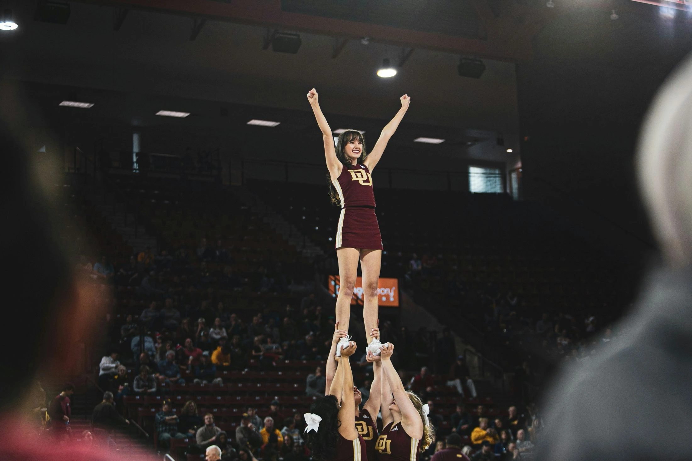 A young female cheerleader with a joyful expression stands atop a pyramid stunt in a dark arena filled with cheering fans.