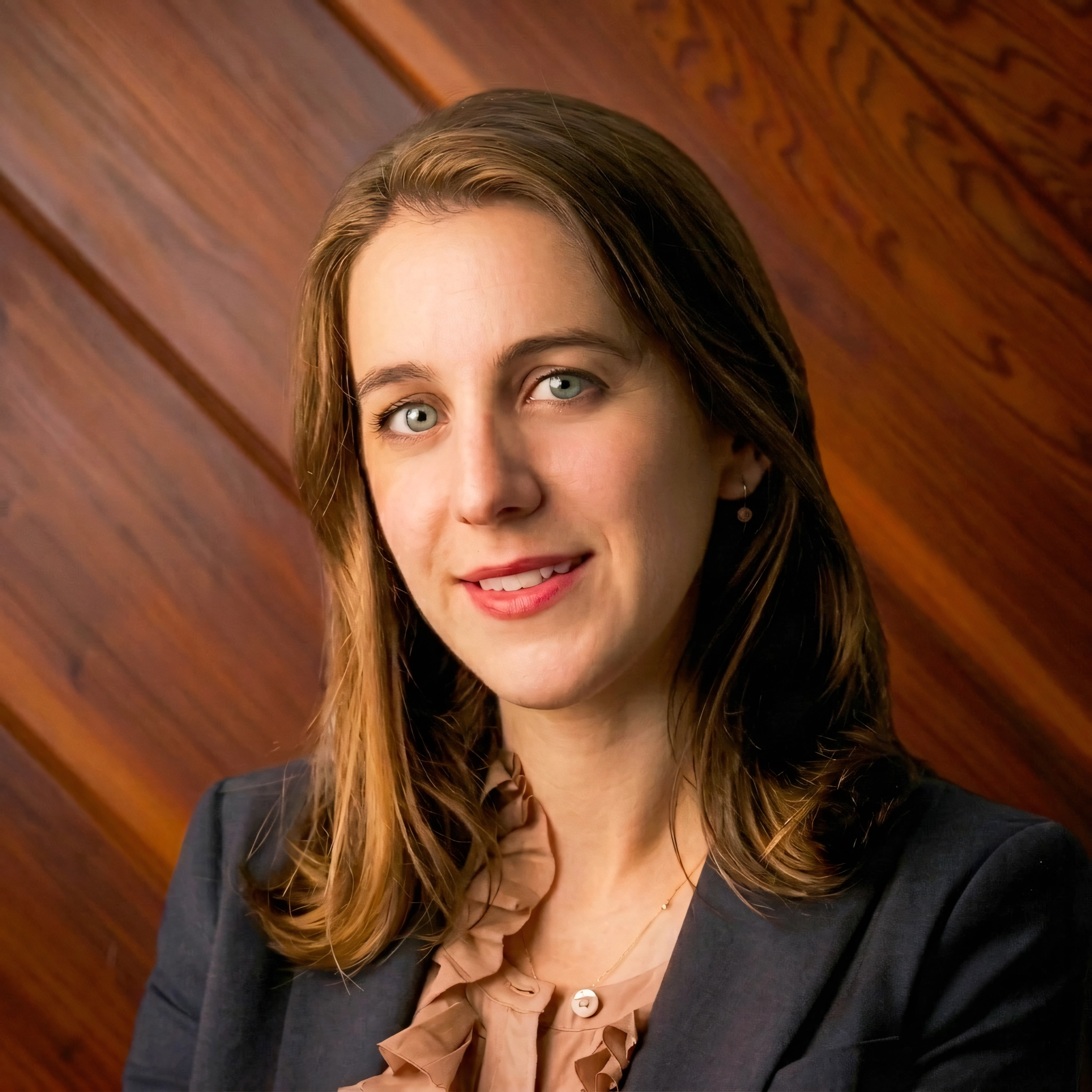 Olympias Iliana Konidaris, an attorney, smiles in a professional headshot. She wears a dark blazer over a ruffled blouse against a warm wood paneled background.