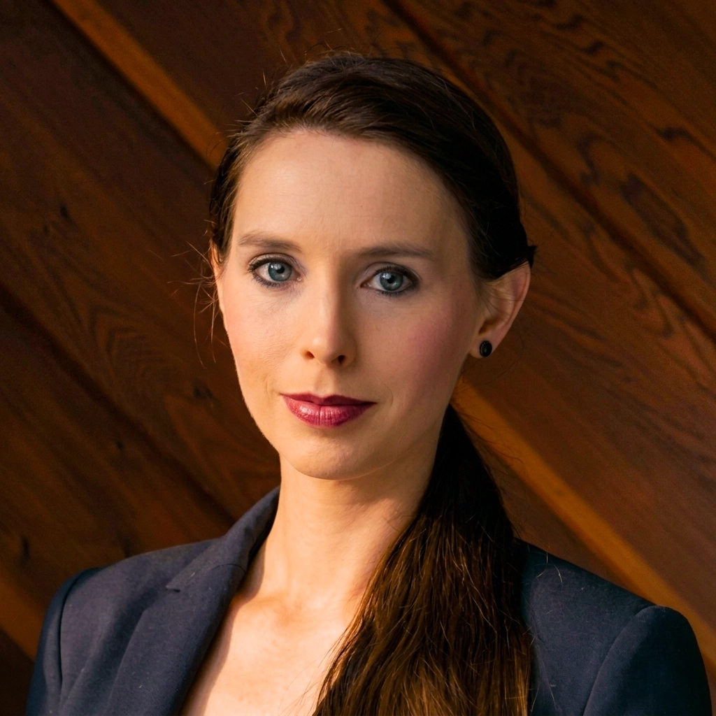 Rachael Denhollander in a dark business blazer, looking forward with a neutral, professional expression. Her dark hair is styled in a sleek ponytail.